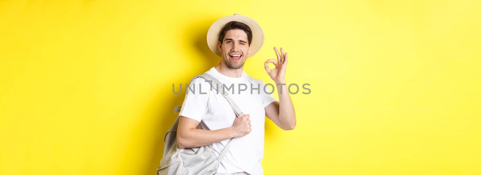 Tourism, travelling and holidays concept. Happy tourist going on vacation, holding backpack and showing okay sign smiling satisfied, standing against yellow background.