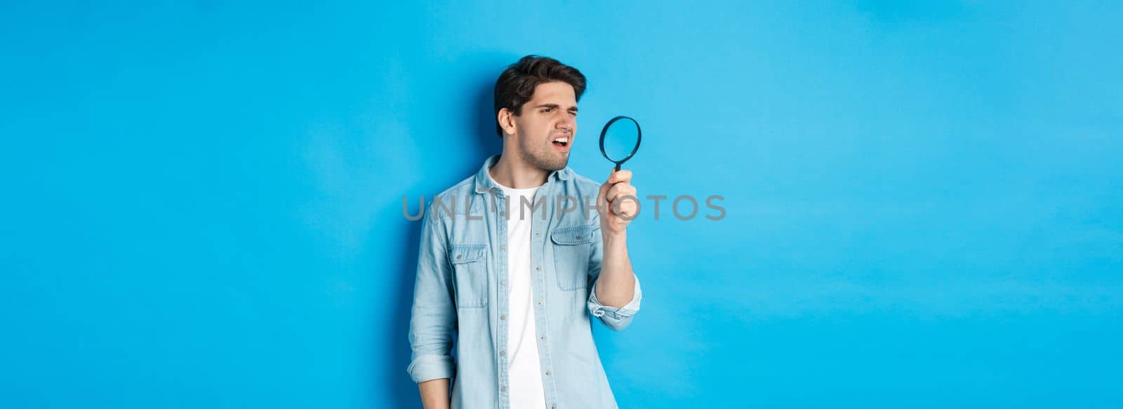 Young thoughtful guy looking through magnifying glass, reading something tiny, standing over blue background.