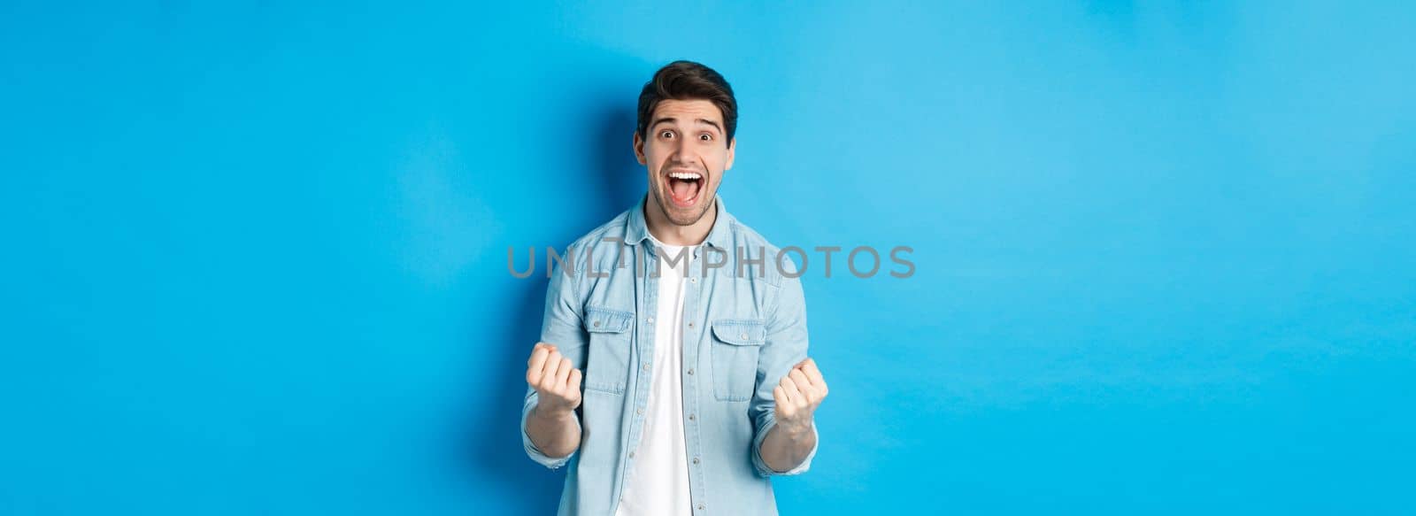 Excited handsome man winning, feeling lucky and rejoicing, celebrating victory and shouting yes, standing against blue background.