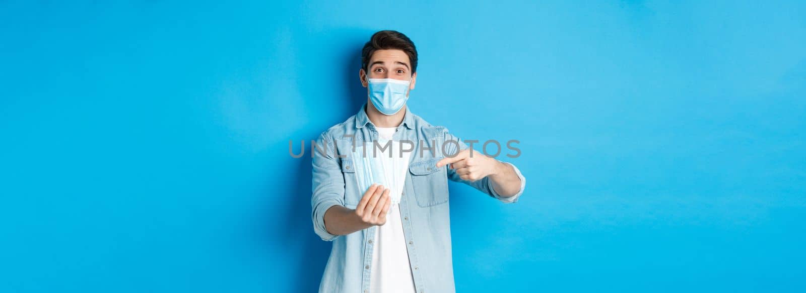 Concept of coronavirus, quarantine and social distancing. Young man pointing at medical masks, preventing measures from covid-19, standing over blue background.