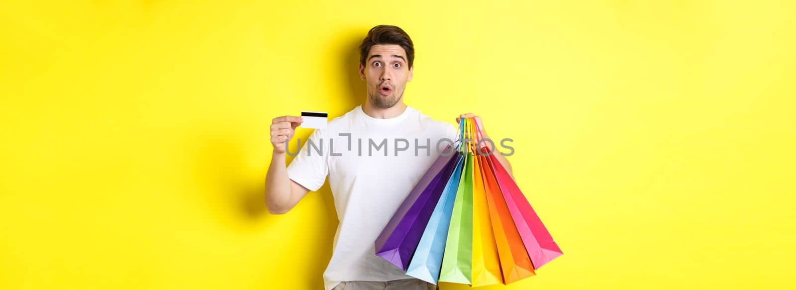 Excited man shopping on black friday, holding paper bags and credit card, standing against yellow background.