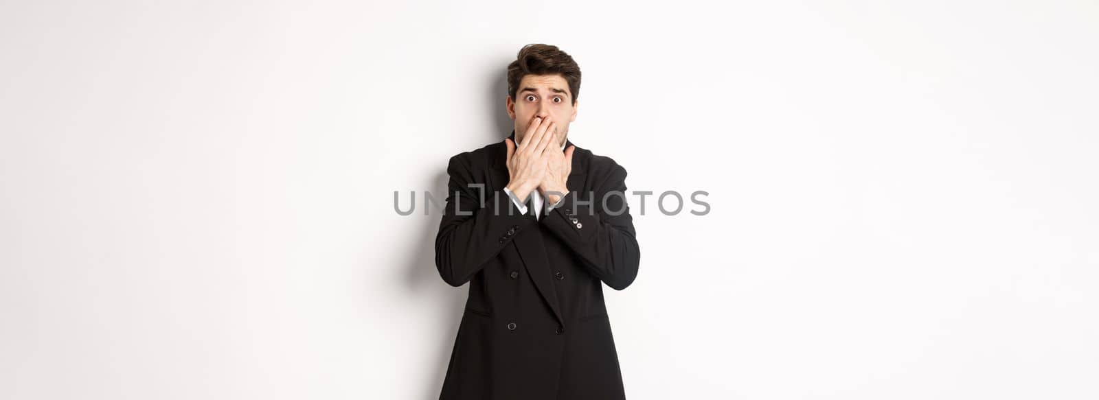 Scared man in formal suit, gasping and looking frightened at camera, standing against white background.