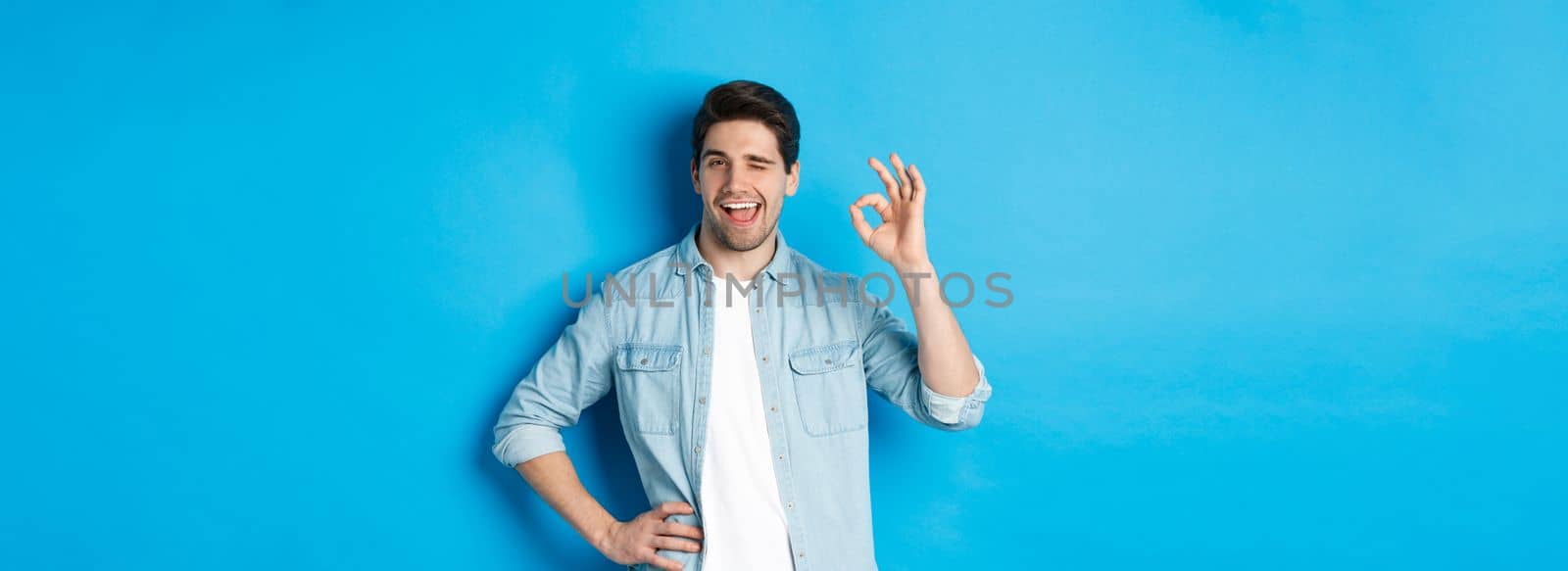 Confident adult man winking and showing okay sign, approve something good, standing against blue background by Benzoix