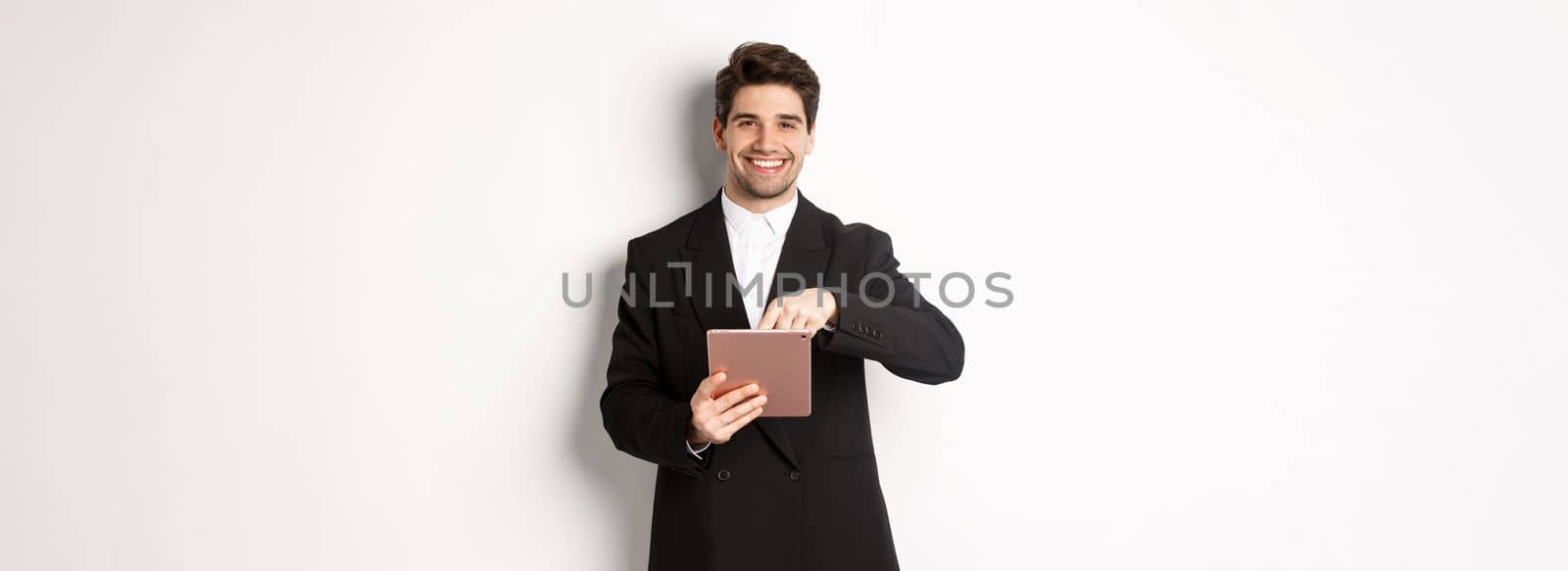 Portrait of handsome, stylish male entrepreneur in black suit pointing at digital tablet, showing something online, standing against white background by Benzoix