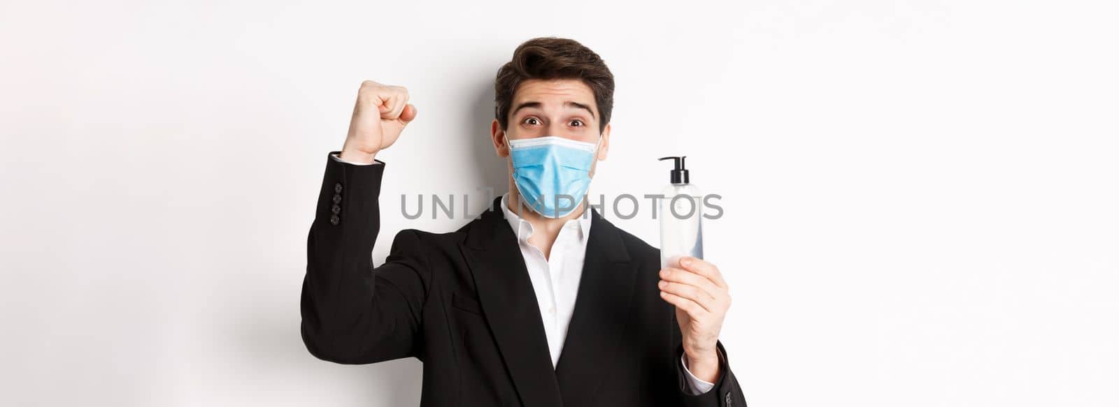 Concept of covid-19, business and social distancing. Close-up of happy man in trendy suit and medical mask, cheering and raising hand up, showing hand sanitizer, white background by Benzoix