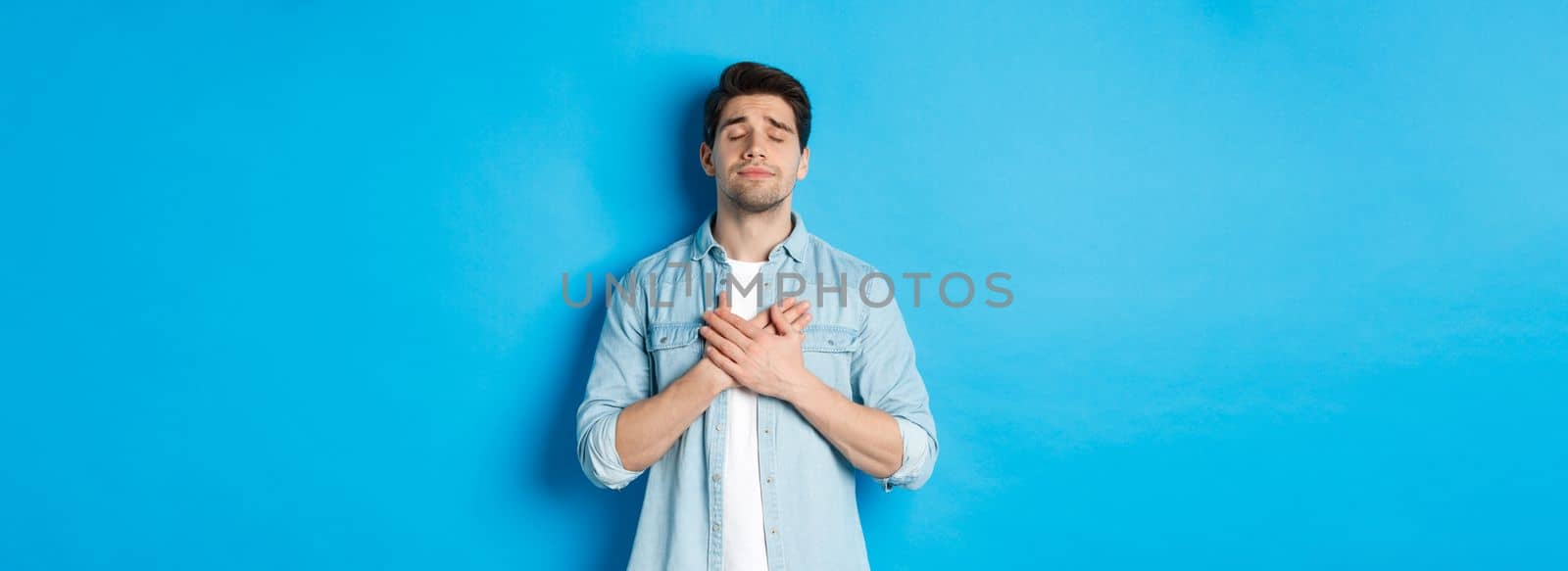 Portrait of dreamy and nostalgic guy in casual outfit, holding hands on heart and close eyes, daydreaming against blue background.