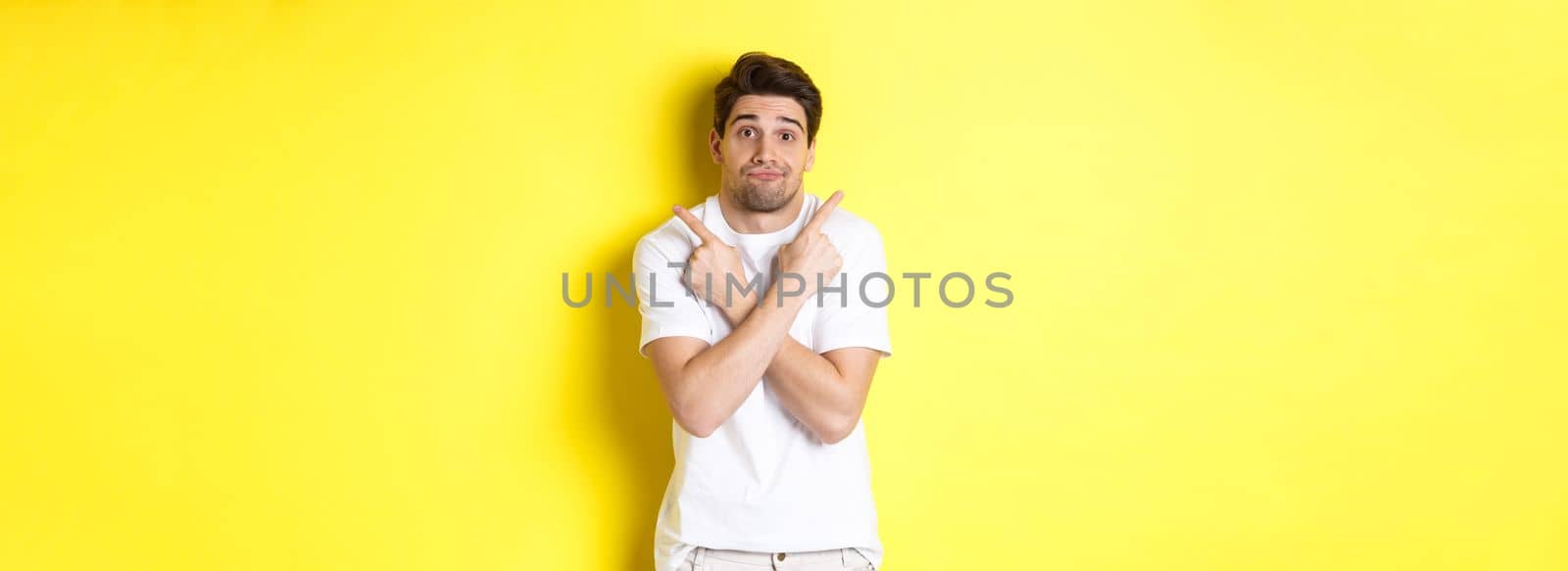 Indecisive man pointing fingers sideways, struggling to make decision, asking advice, standing over yellow background.