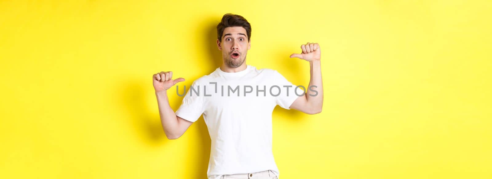 Surprised handsome guy pointing at himself, looking amazed, standing over yellow background by Benzoix