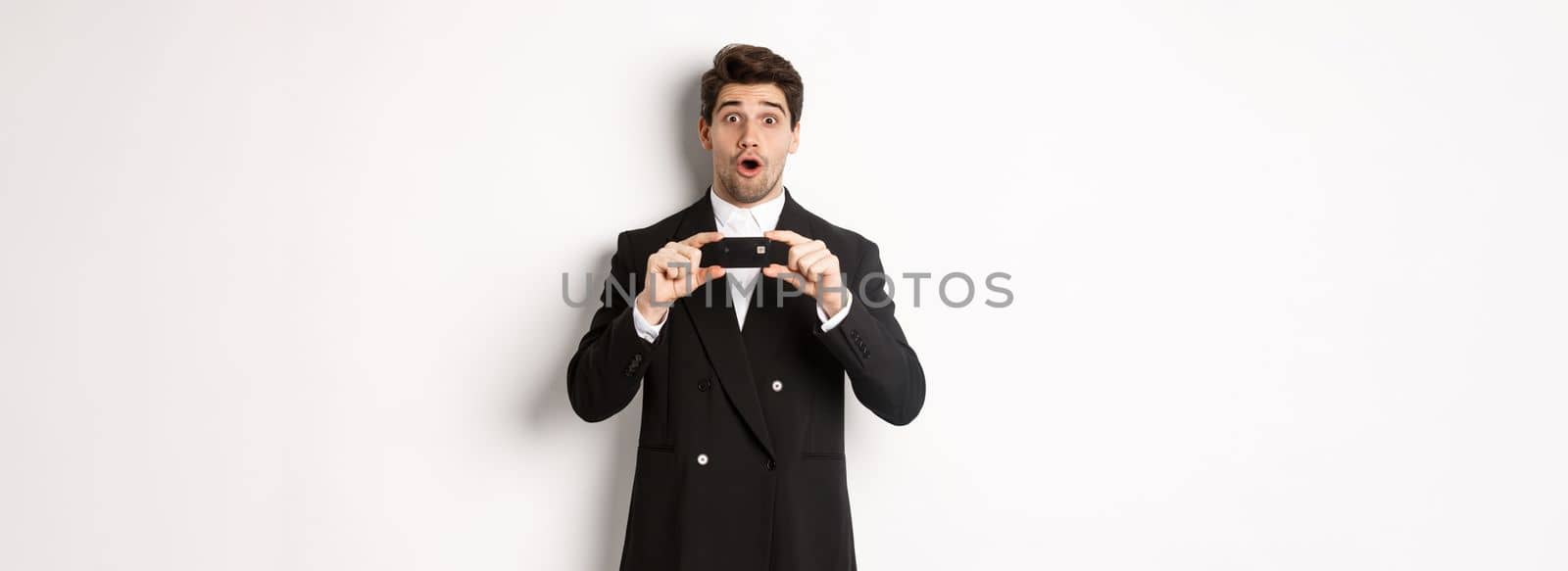 Portrait of handsome businessman in black suit, showing credit card and looking amazed, standing over white background.