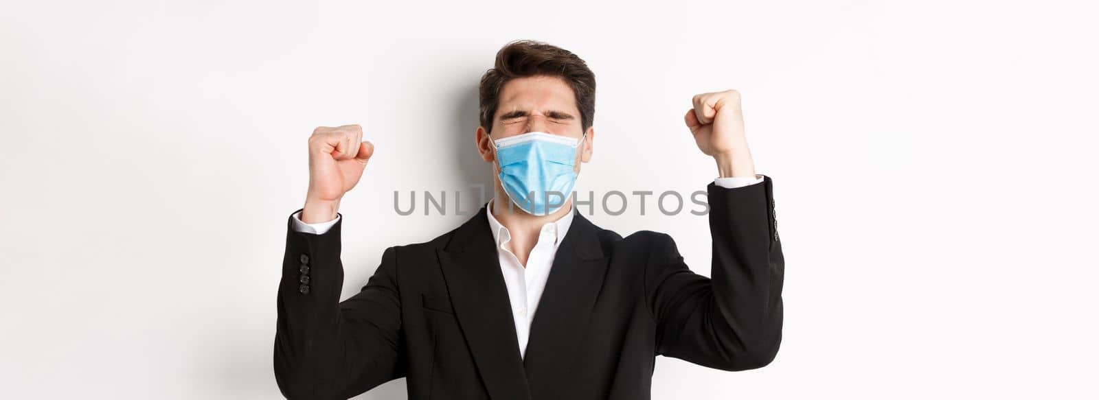 Concept of covid-19, business and social distancing. Close-up of handsome man in suit and medical mask, rejoicing and winning, raising hands up, shouting yes.