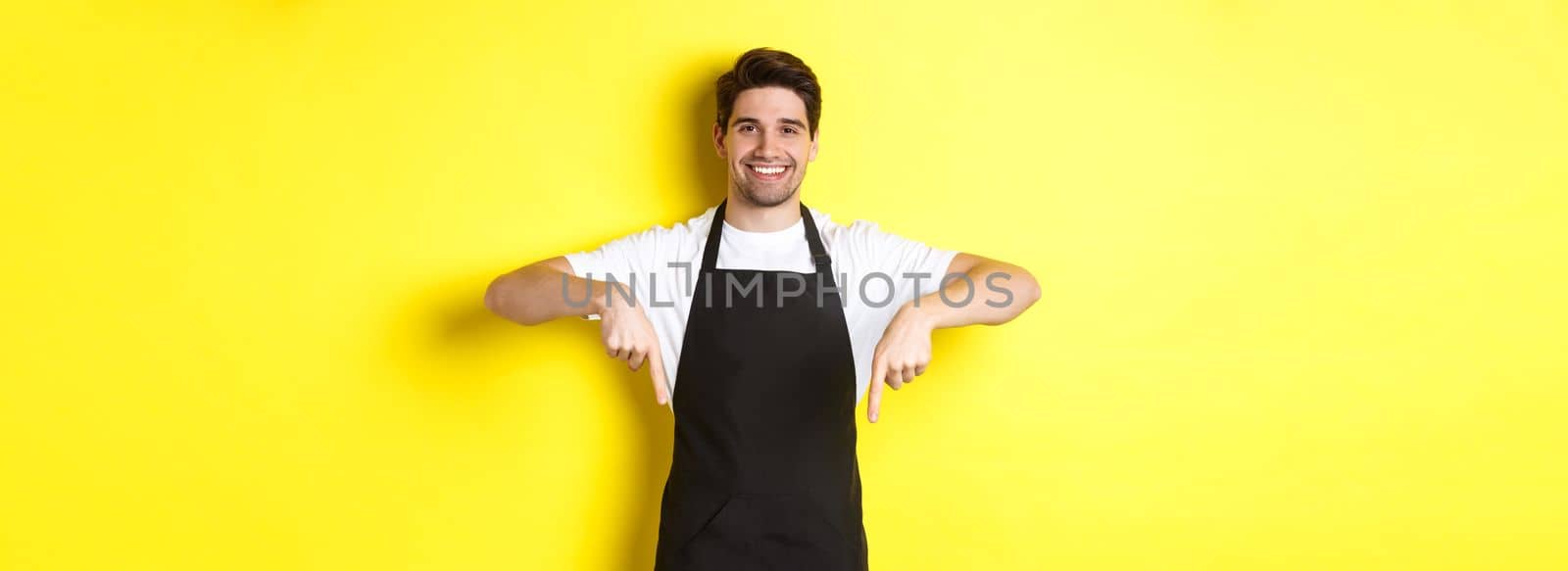 Friendly barista in black apron pointing fingers down, showing your logo banner, standing over yellow background.