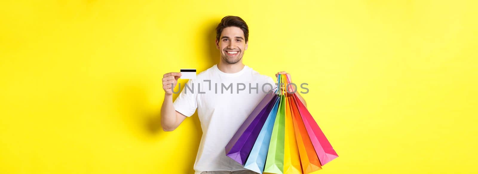 Young man shopping for holidays, holding paper bags and recommending bank credit card, standing over yellow background.