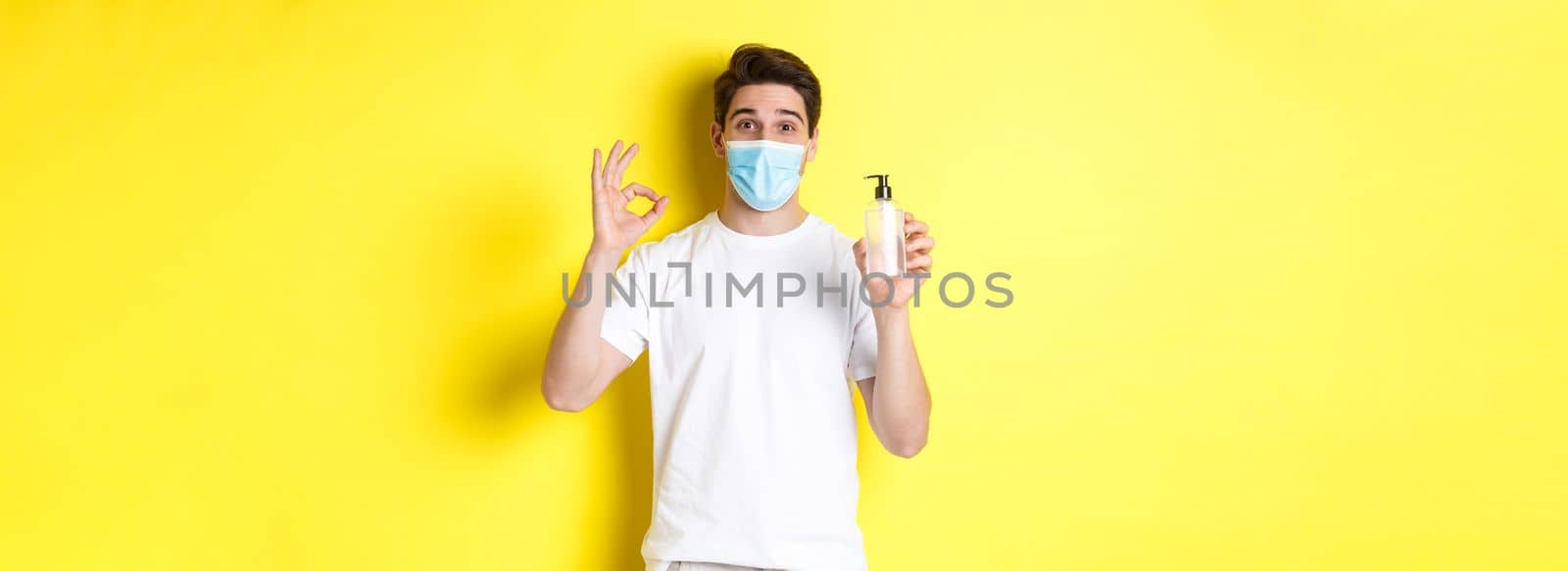 Concept of covid-19, quarantine and lifestyle. Satisfied young man in medical mask showing good hand sanitizer, make okay sign and recommending antiseptic, yellow background.