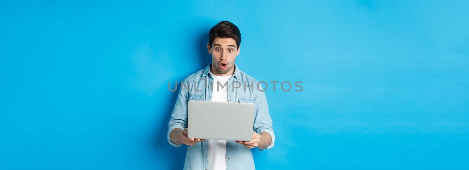 Impressed caucasian guy looking at laptop screen with amazement, checking out promo in internet, standing against blue background.