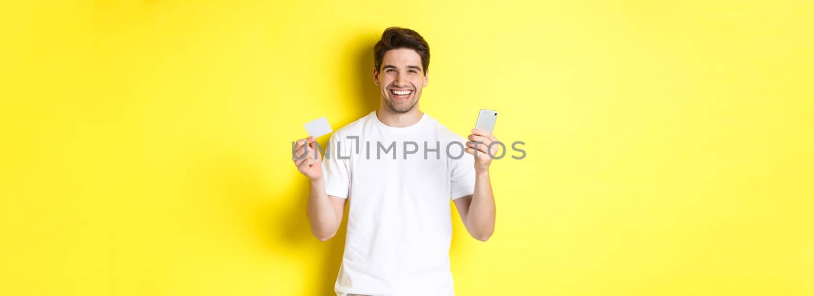 Happy young man shopping online in smartphone, holding credit card and smiling, standing over yellow background.