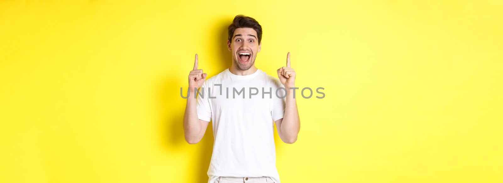 Portrait of excited handsome man in white t-shirt, pointing fingers up, showing offer, standing against yellow background.