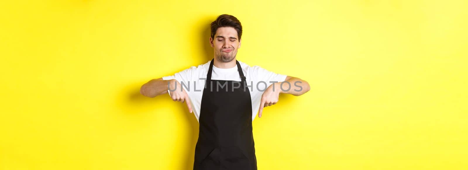 Doubtful barista complaining, pointing fingers down and grimacing displeased, standing in black apron against yellow background.