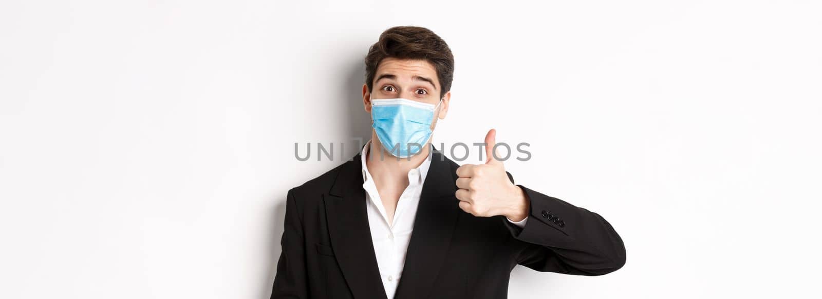 Concept of covid-19, business and social distancing. Close-up of happy businessman in black suit and medical mask, showing thumbs-up, making a compliment, white background.