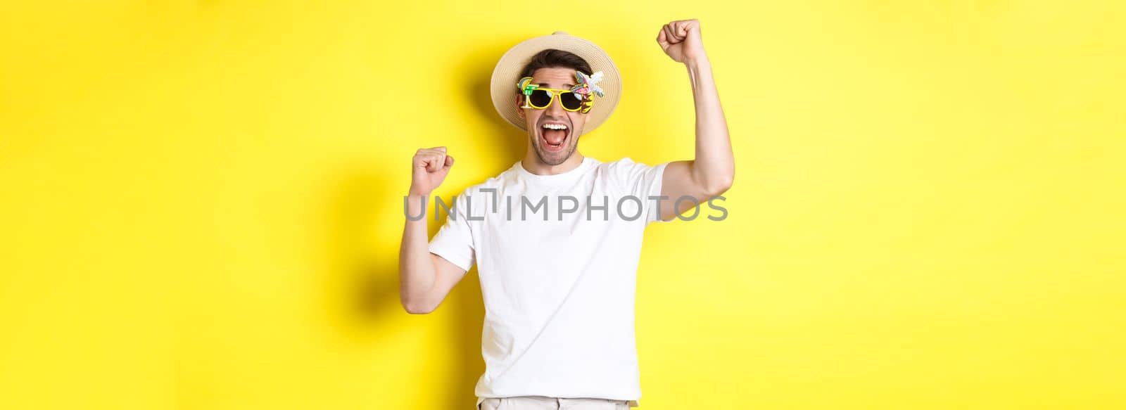 Concept of tourism and lifestyle. Happy man winning trip to resort, shouting yes and raising hands up, triumphing, wearing sunglasses and summer hat, yellow background by Benzoix