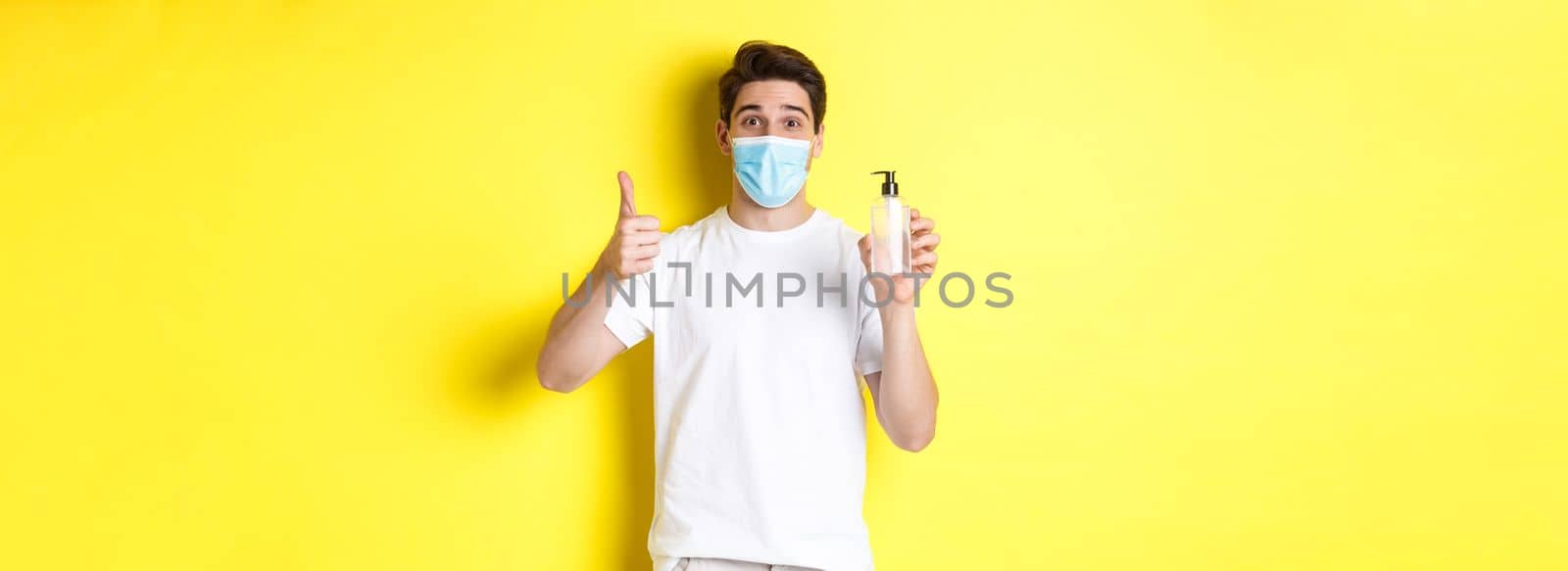 Concept of covid-19, quarantine and lifestyle. Satisfied young man in medical mask showing good hand sanitizer, thumbs up and recommending antiseptic, yellow background.