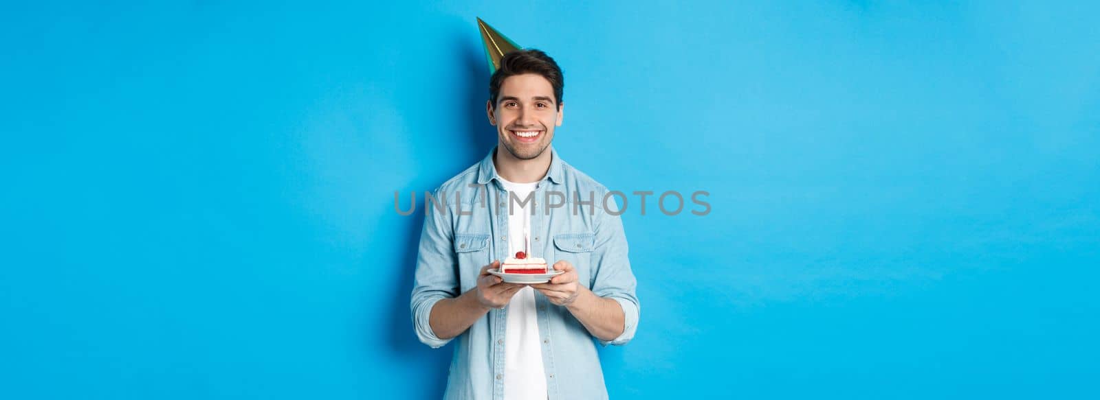Smiling man holding b-day cake and wearing birthday party hat, celebrating over blue background.