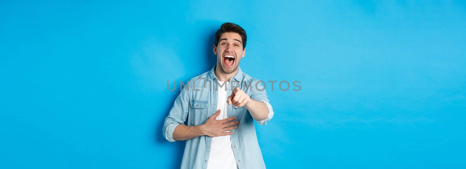 Adult man in casual outfit laughing out loud and pointing at you, looking at something funny, chuckling while standing against blue background.