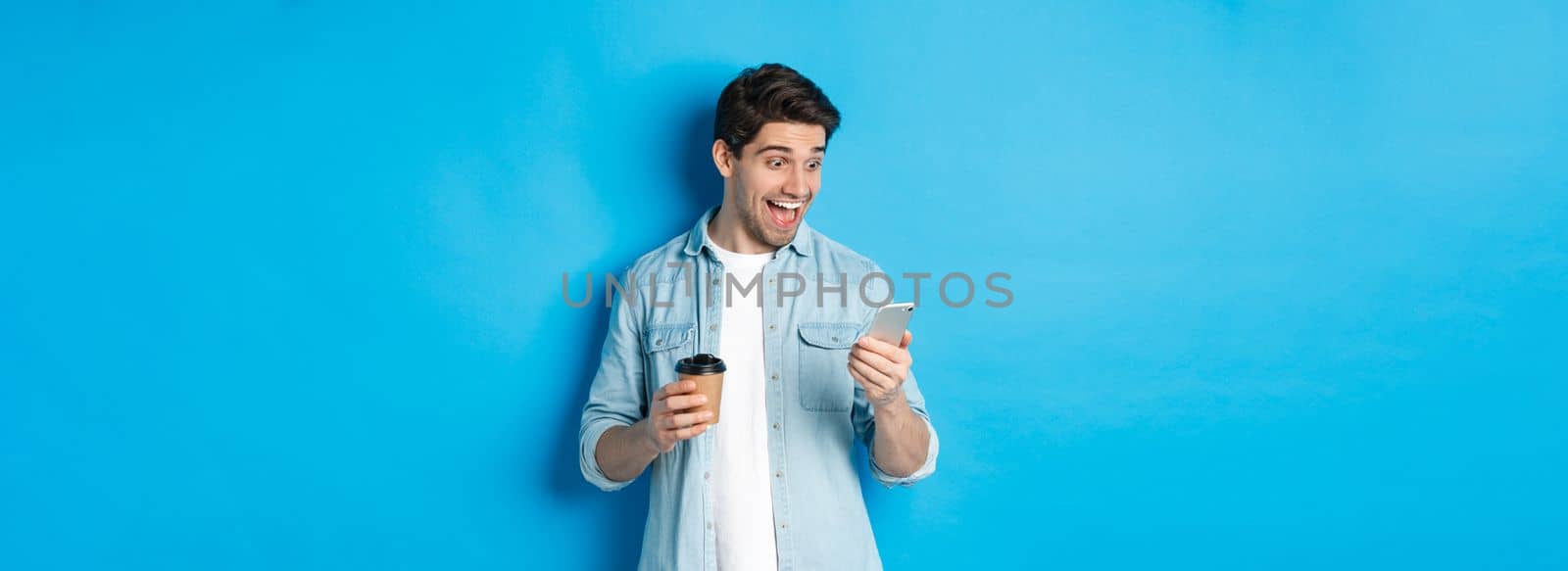 Man looking surprised at smartphone, drinking coffee, standing over blue background amazed.