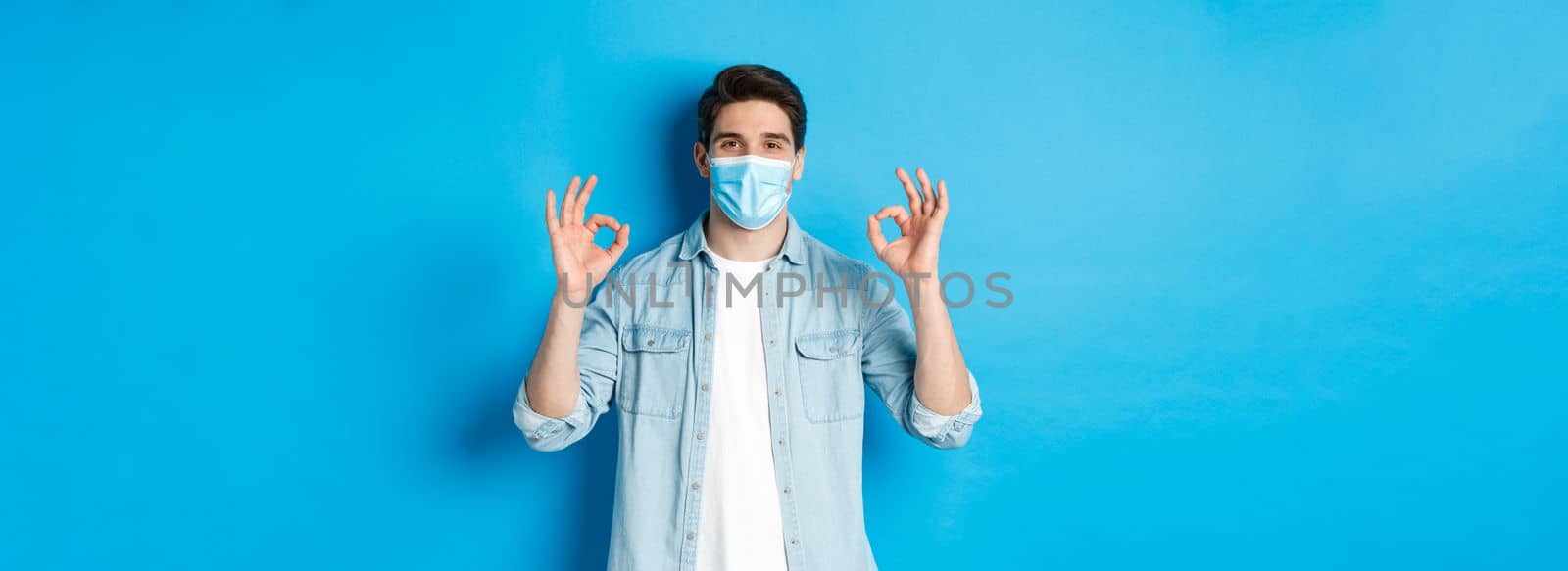 Concept of covid-19, pandemic and social distancing. Satisfied man in medical mask, showing ok signs in approval, agree or like something good, standing over blue background.