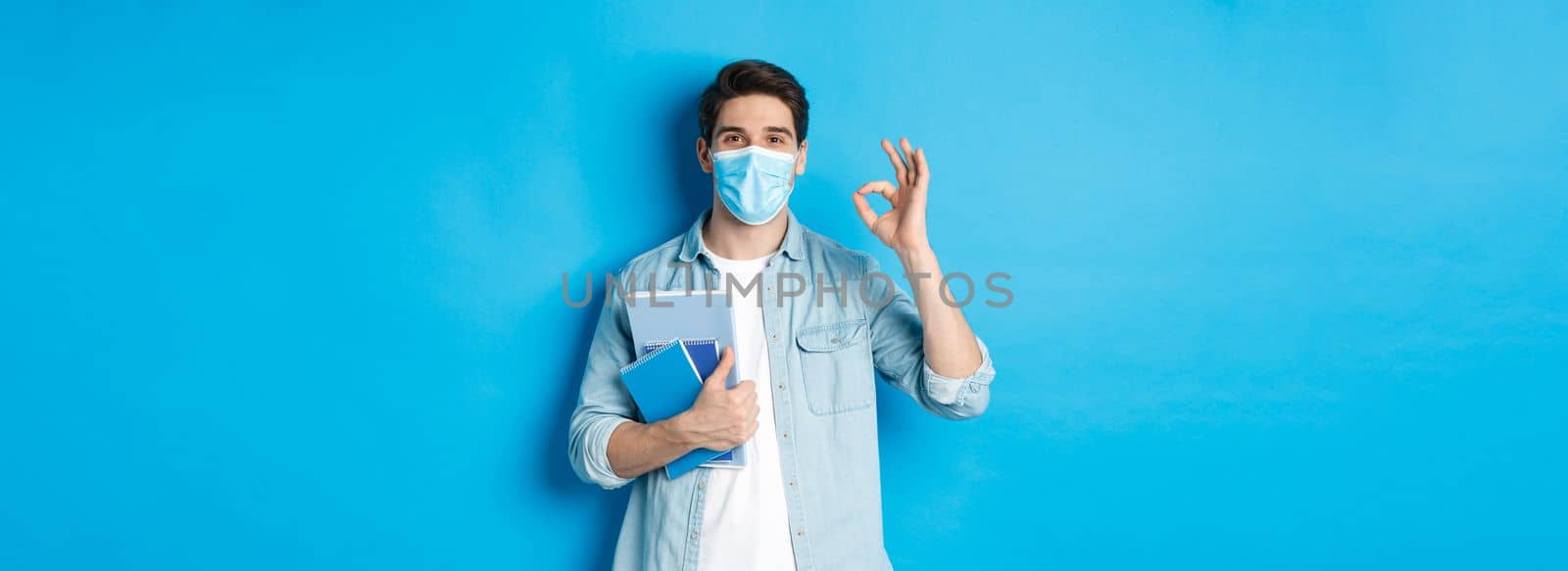 Education, covid-19 and social distancing. Guy student in medical mask looking happy, holding notebooks, showing ok sign, standing over blue background.