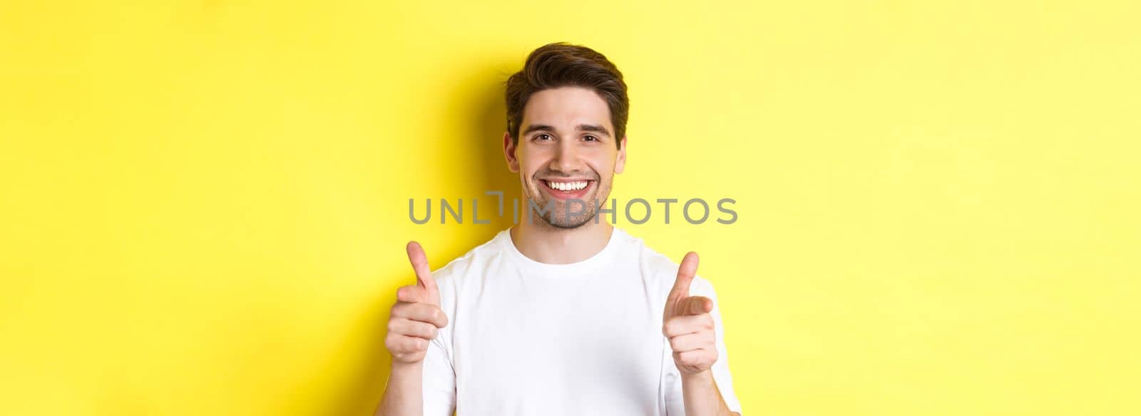 Confident man pointing fingers at camera and smiling, praising you, standing over yellow background.