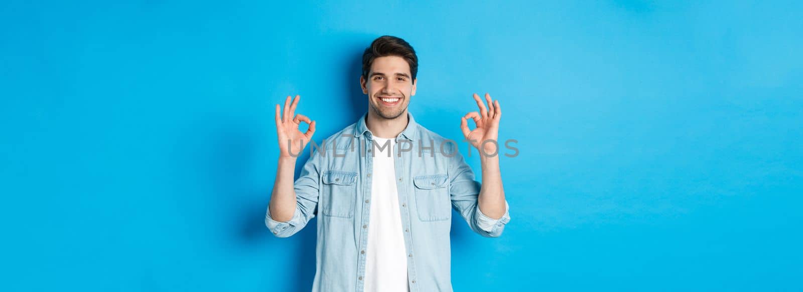 Confident smiling adult man, showing ok signs and looking pleased, like something, standing against blue background by Benzoix