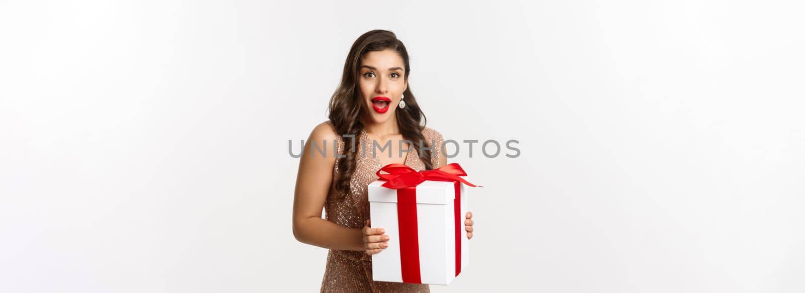 Merry Christmas. Image of beautiful woman in glamour dress receiving gift and looking surprised, celebrating New Year, standing over white background.