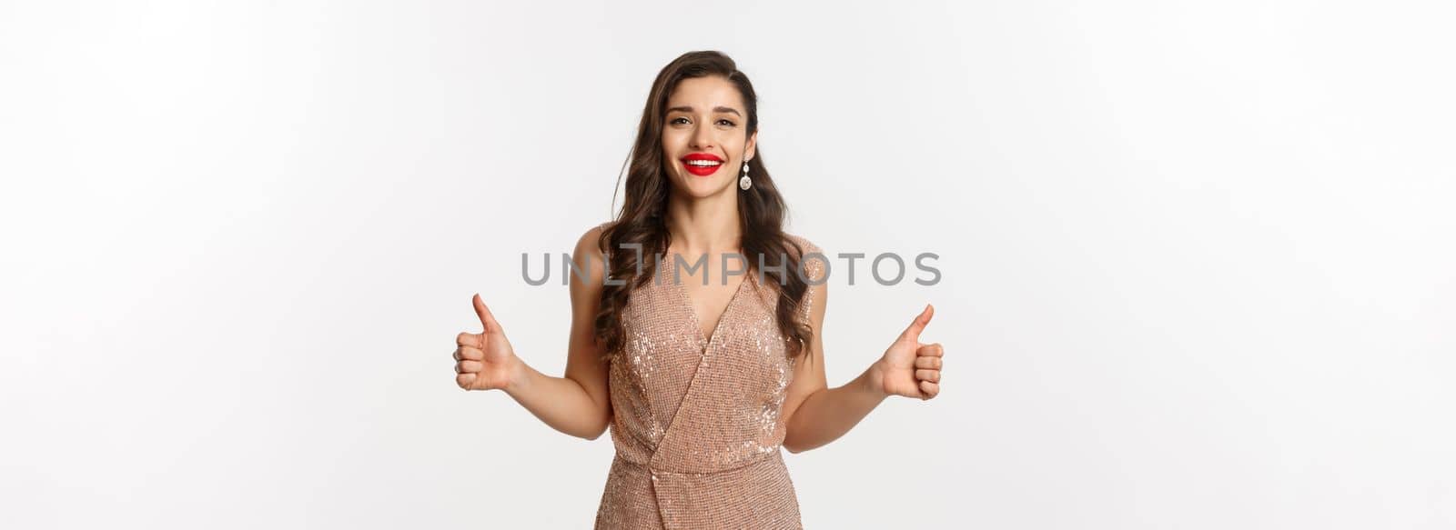 Concept of celebration, holidays and party.Attractive female model in luxurious dress, showing thumbs up and smiling satisfied, approve and like, standing over white background.