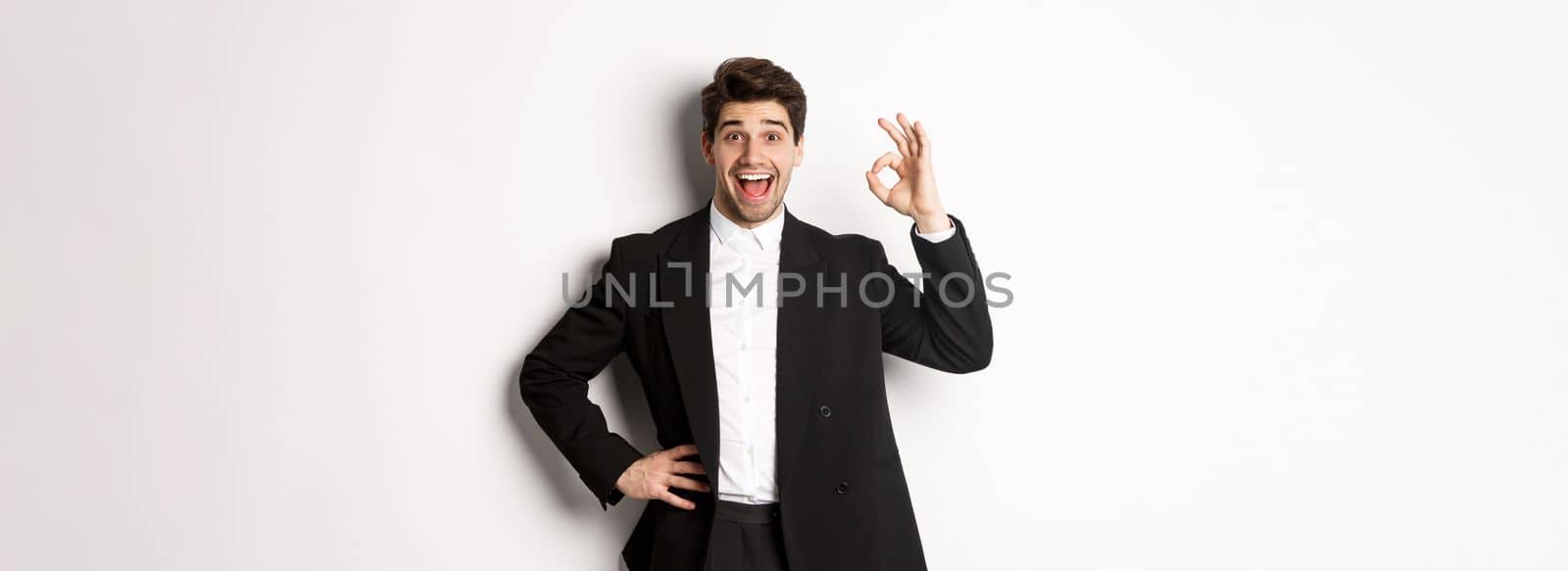 Concept of new year party, celebration and lifestyle. Portrait of pleased handsome guy in black suit, praise something good, showing okay sign in approval, standing over white background by Benzoix