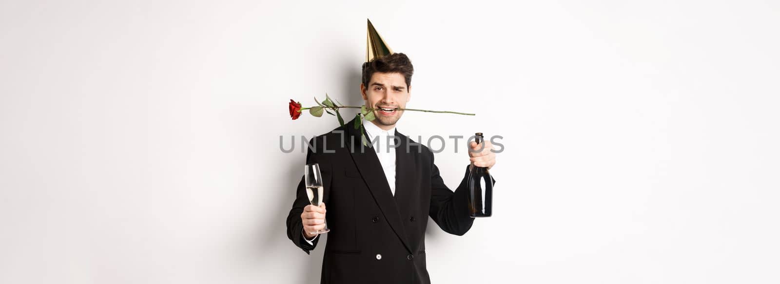 Romantic guy in trendy suit, celebrating and having a party, holding rose in teeth and champagne, standing over white background by Benzoix