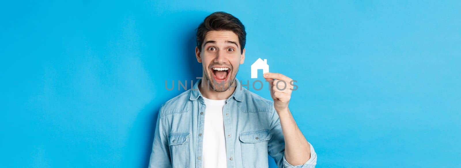 Real estate concept. Happy young man looking excited, found apartment, showing small house model, standing over blue background.