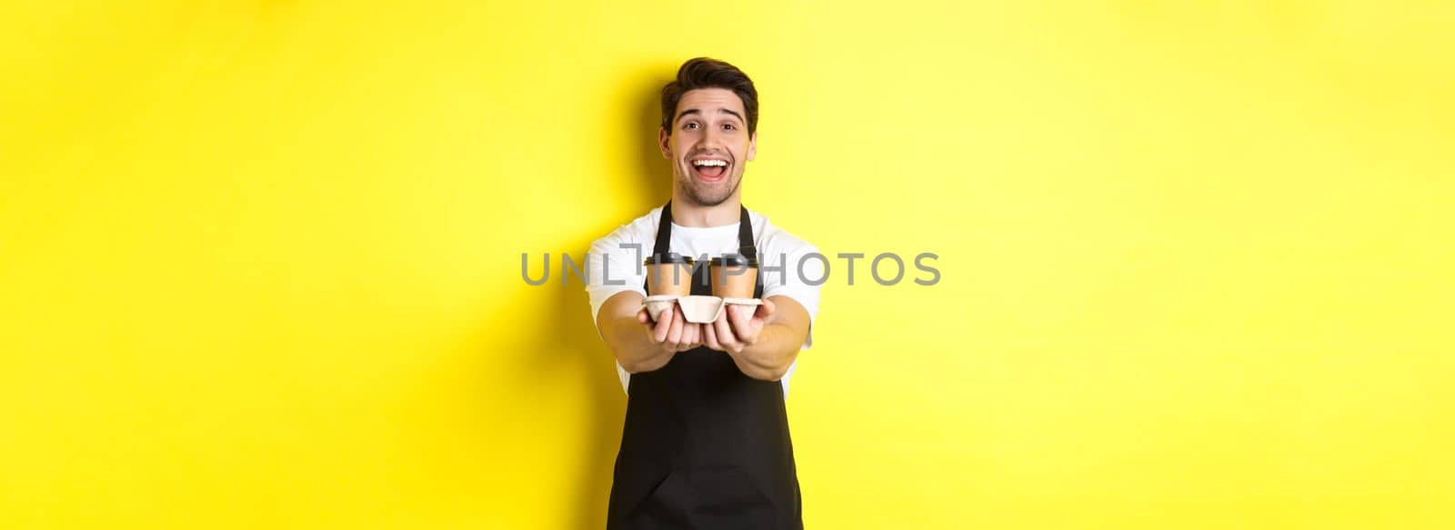 Friendly barista in black apron giving takeaway order, holding two cups of coffee and smiling, standing over yellow background.