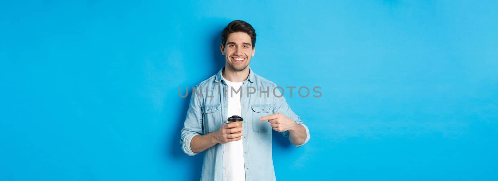 Smiling happy man pointing at paper cup with coffee, recommending cafe, standing over blue background.