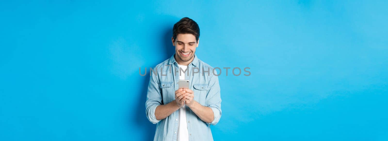 Image of handsome young man using mobile phone, texting on phone and looking pleased, standing over blue background by Benzoix