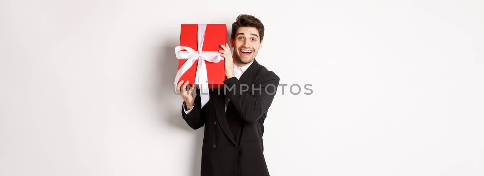 Concept of christmas holidays, celebration and lifestyle. Attractive man in black suit, holding new year gift and smiling, standing with a present over white background.