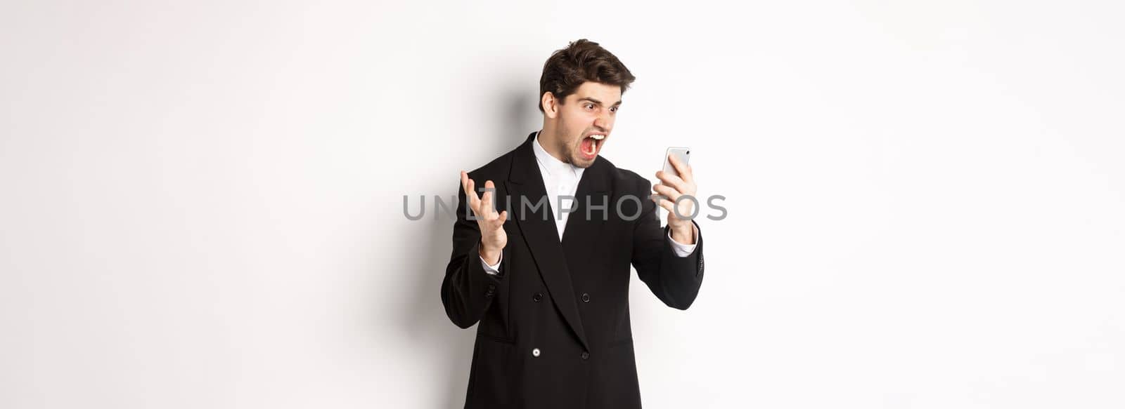 Portrait of angry businessman in black suit yelling at mobile phone, having an argument on video call, standing mad over white background.