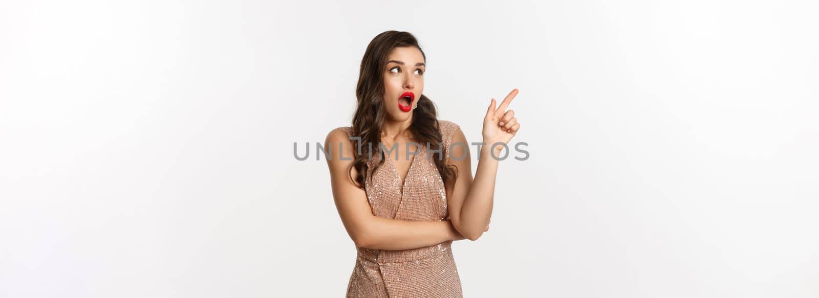 Christmas, holidays and celebration concept. Amazed fashionable woman in party dress and red lipstick, pointing and looking left at promo offer, standing against white background.
