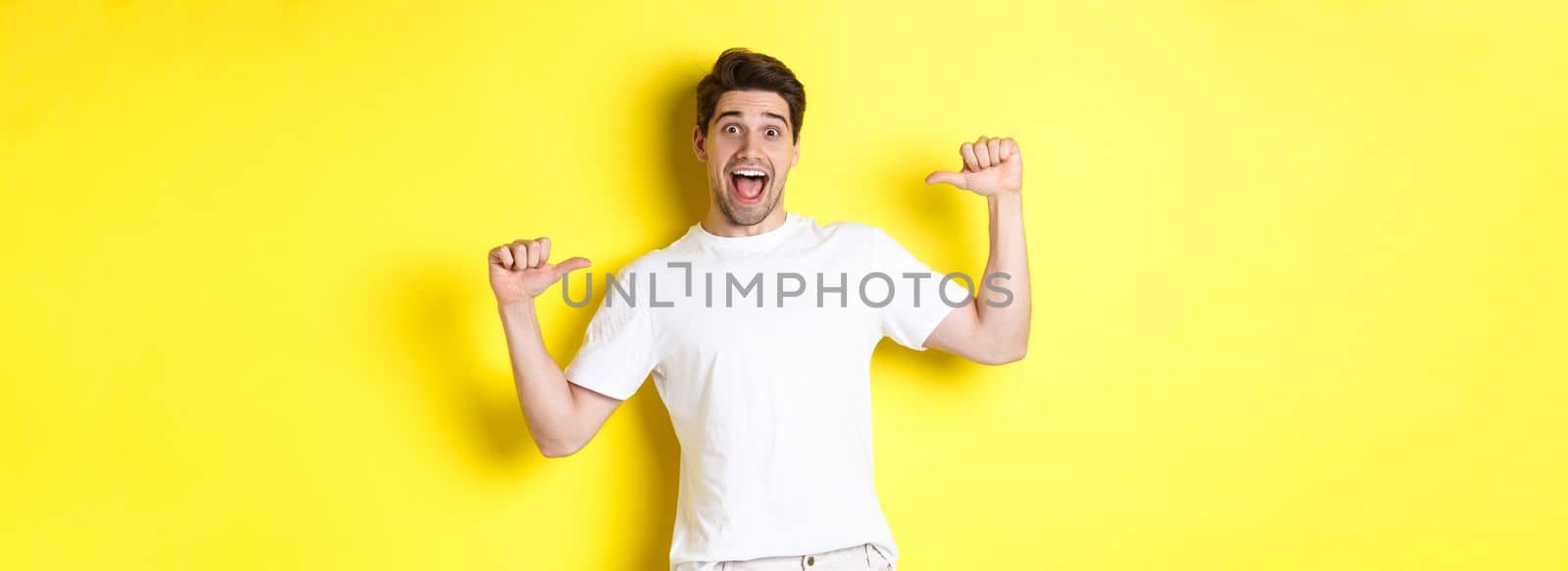 Excited man looking happy, pointing at himself with amazement, standing over yellow background.