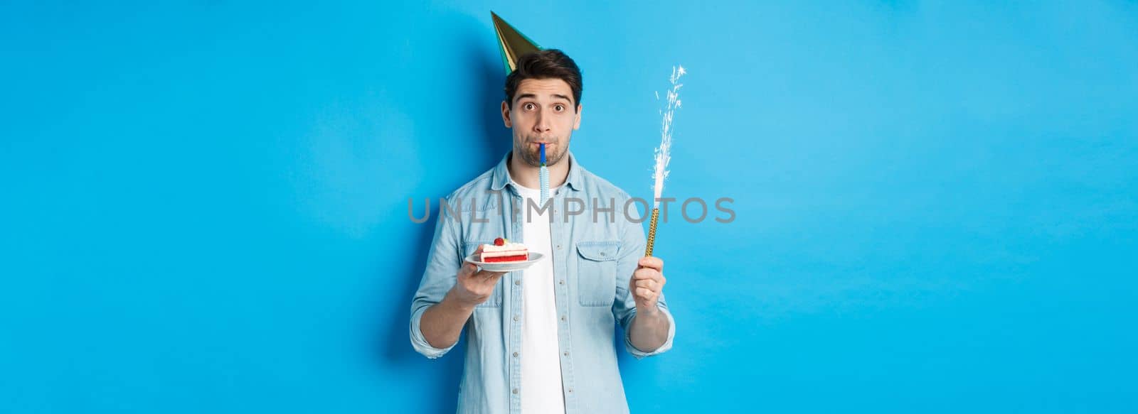 Funny guy celebrating birthday, holding b-day cake, firework and wearing party hat, standing over blue background.