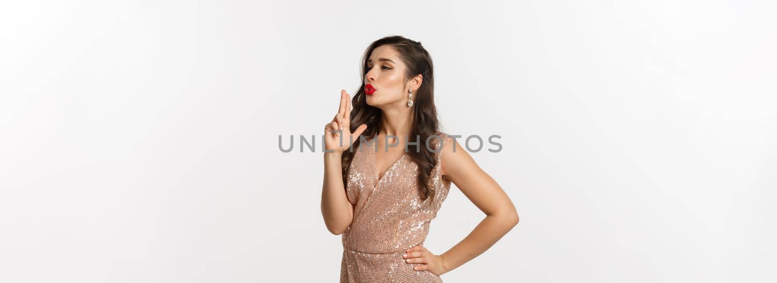 Concept of casino, celebration and party. Beautiful female model with red lips, wearing glamour dress, blowing at finger gun after making shot, standing over white background.