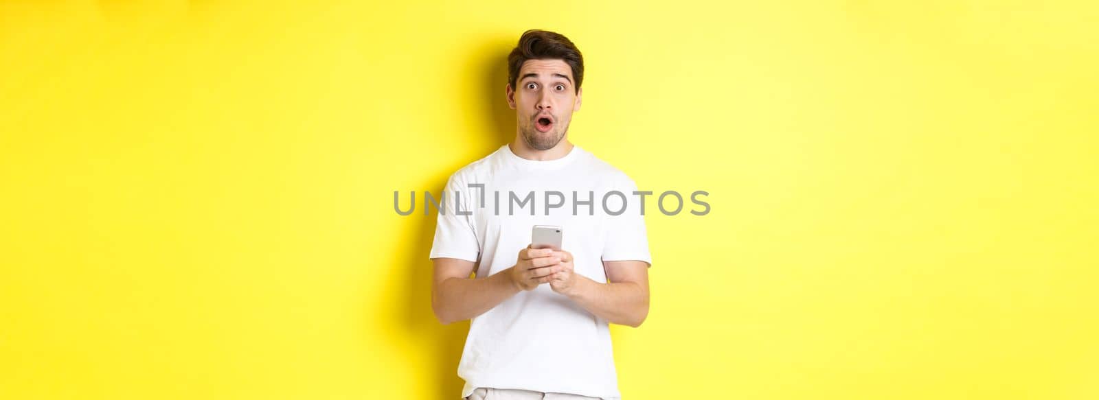 Man looking surprised, using smartphone, open mouth and saying wow, standing against yellow background by Benzoix