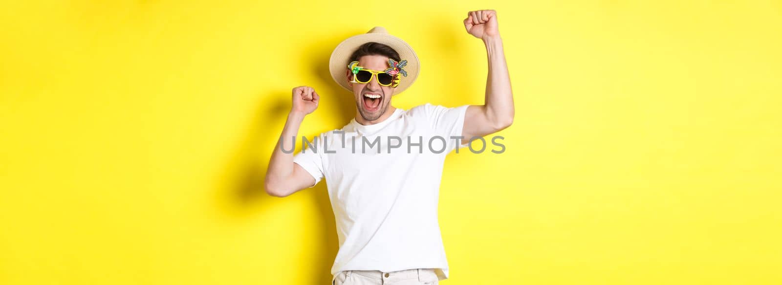 Concept of tourism and lifestyle. Happy lucky guy winning trip, rejoicing and wearing holiday outfit, summer hat and sunglasses, yellow background.