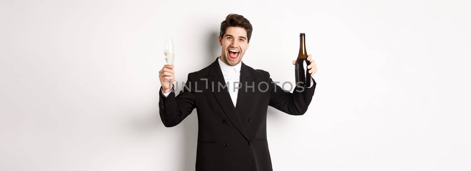 Concept of holidays, party and celebration. Handsome man in trendy suit having fun, holding bottle and glass of champagne, standing over white background.