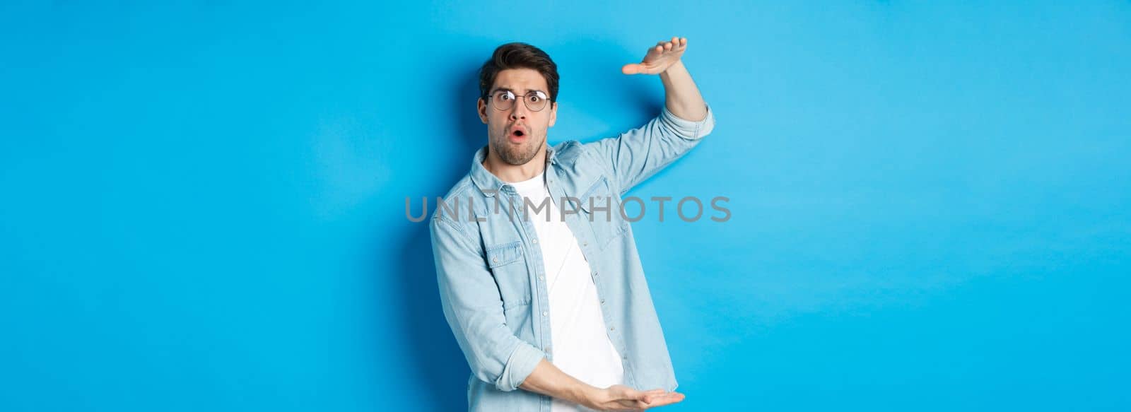 Confused handsome man showing big size, shaping large box and looking amazed, standing over blue background.