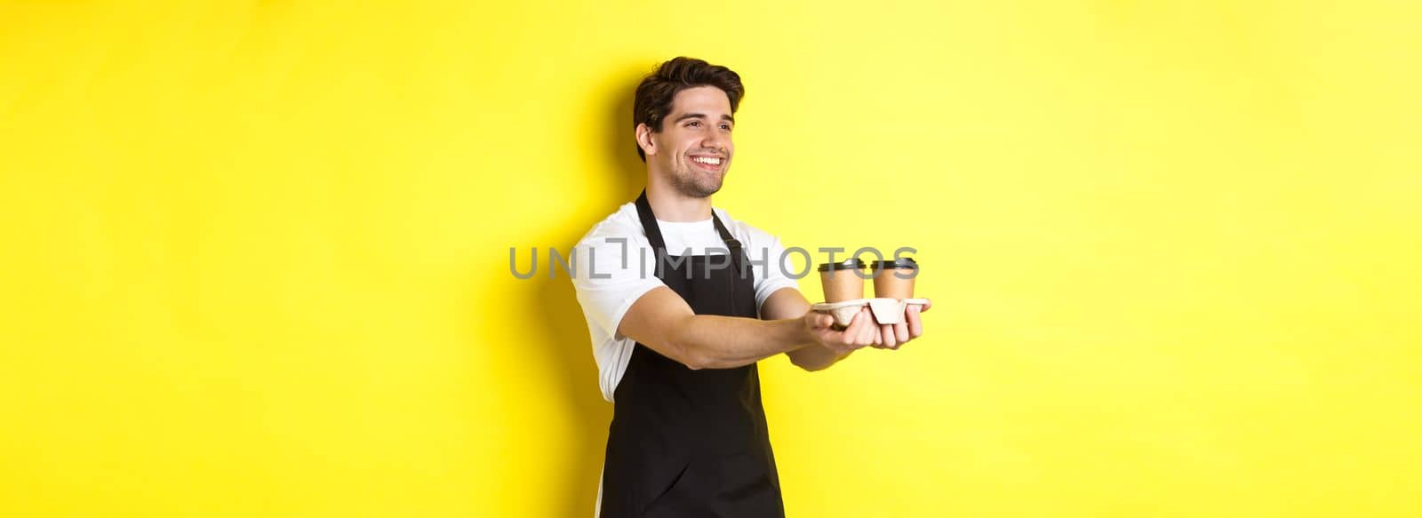 Friendly barista in black apron giving takeaway order, holding two cups of coffee and smiling, standing over yellow background by Benzoix